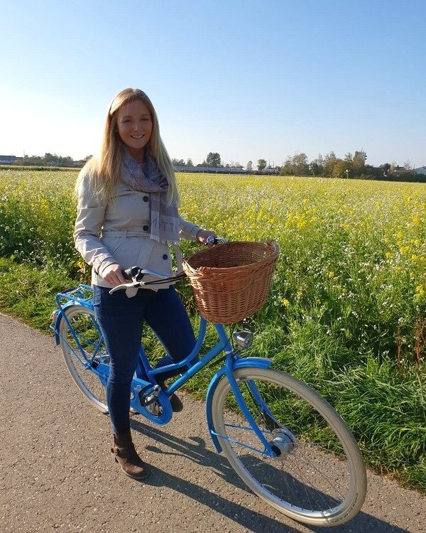 Hollandrad in hellblau, blonde frau, gefolchtener weiden fahrradkorb in hellbraun, helle creme schwalbe reifen road cruiser, rapsfeld im hintergrund